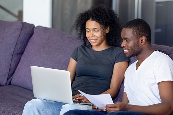 African-married-couple-sitting-on-sofa-at-home-use-laptop-holds-paper-check-bills